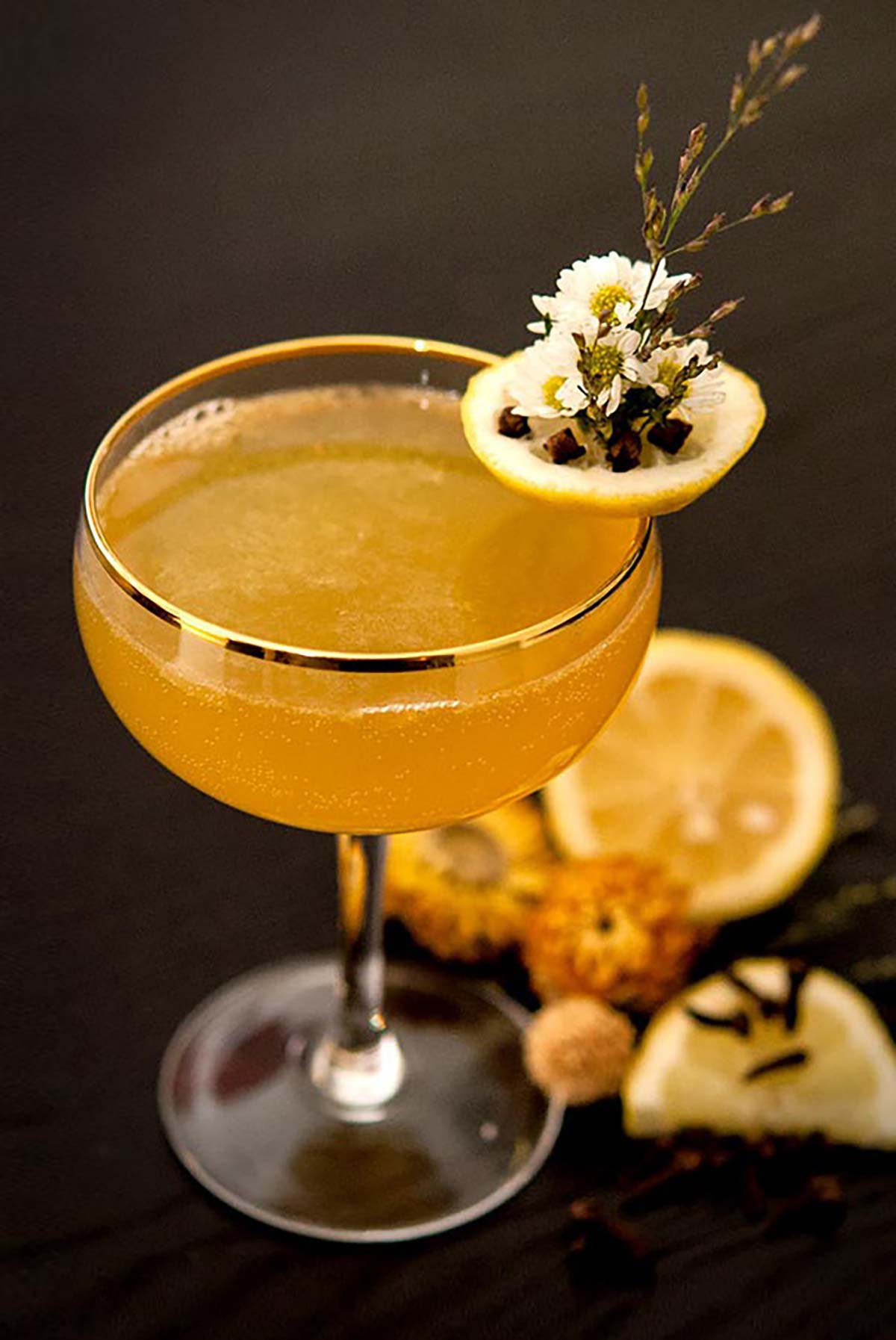 A cocktail garnished with daisies, lemon and cloves, sitting on a black table with sliced lemons and dry flowers around it.