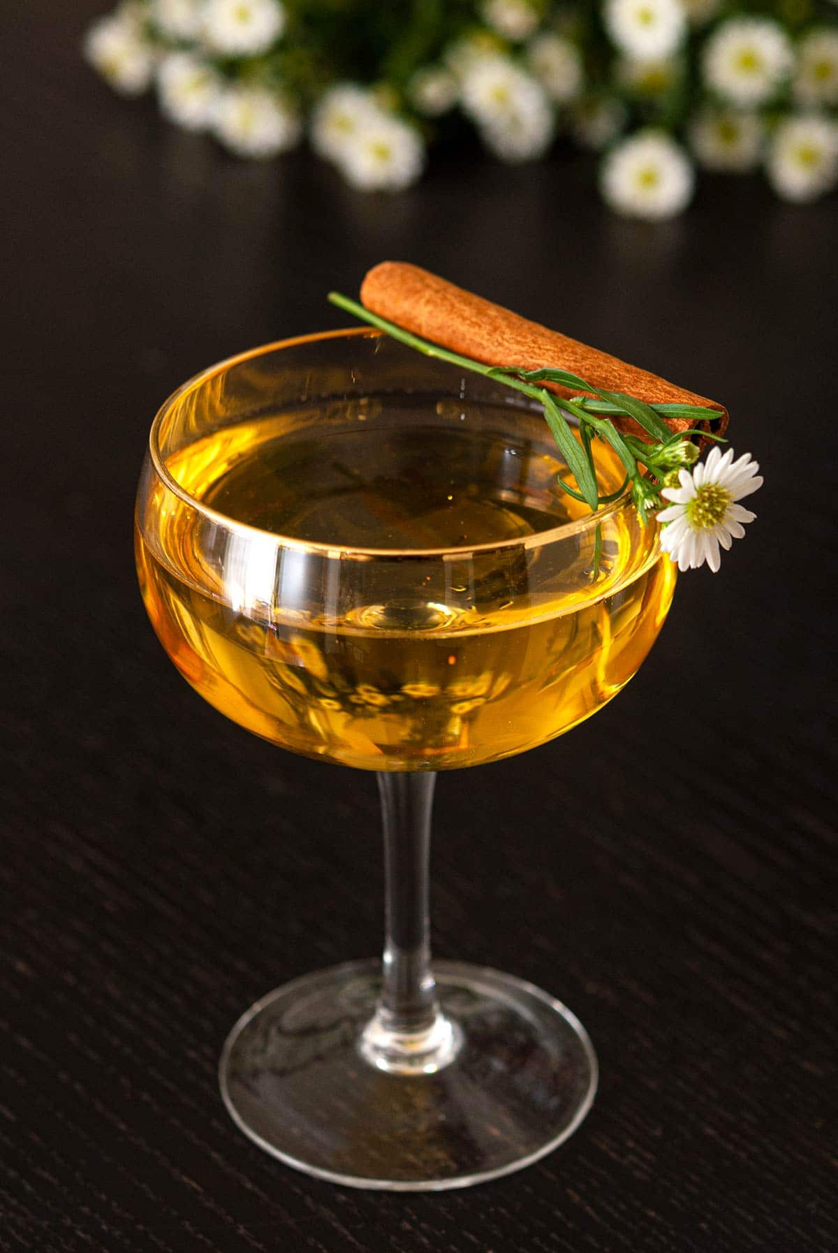 A cocktail with a gold rim on a black table, garnished with a cinnamon stick and daisy.