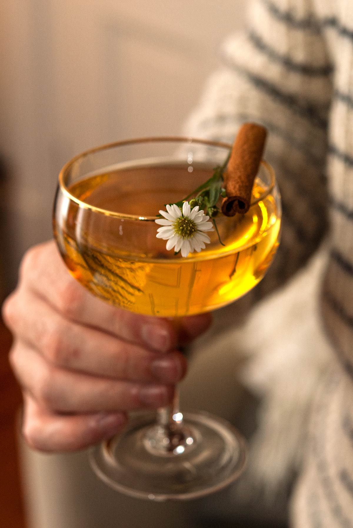 A man's hand holding a cocktail garnished with a daisy and cinnamon stick.