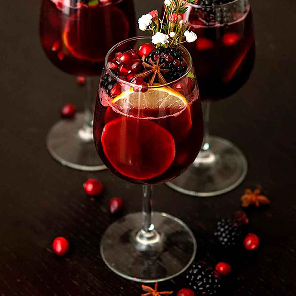 A glass of sangria garnished with fruits, flowers, herbs and star anise, with a few scattered cranberries and blackberries.