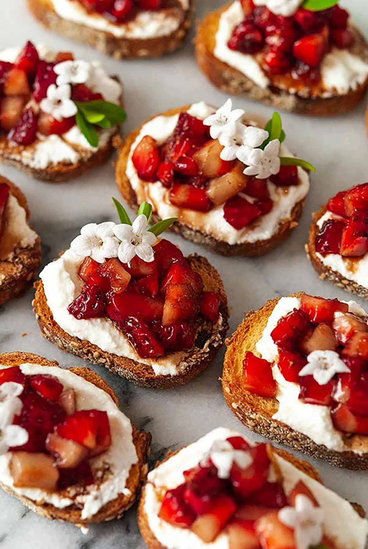 10 balsamic strawberry crostini, garnished with small flowers, on a marble plate.