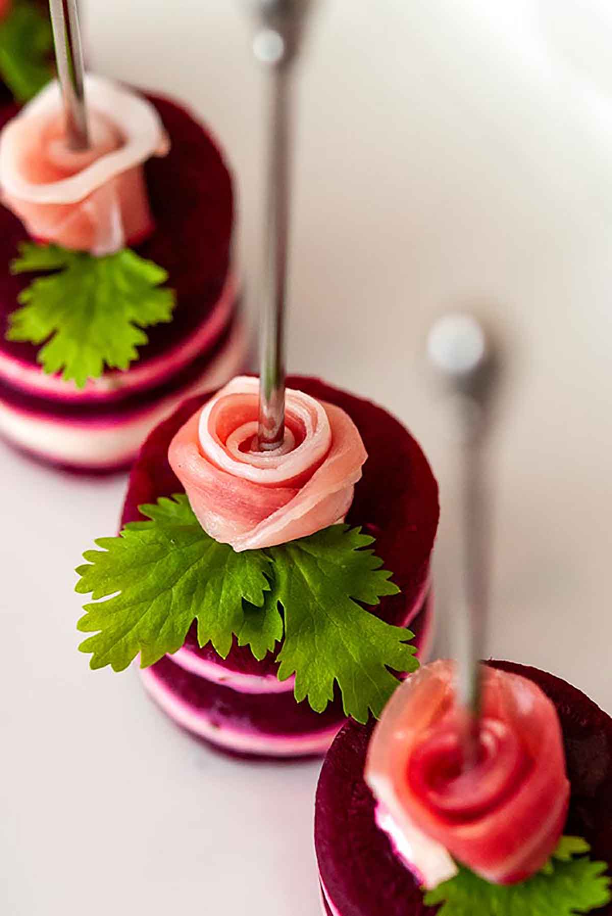 3 small beet napoleons, with a prosciutto rose and cilantro leaf on top, with cocktail pins through the center of the roses.