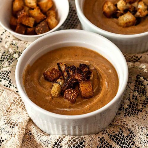 2 bowls of cauliflower soup and a small bowl of croutons on a lace table cloth.