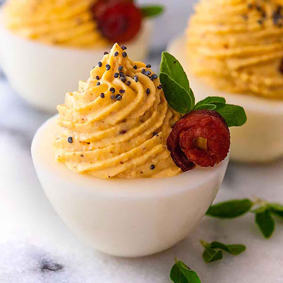 3 deviled eggs garnished with tiny bacon roses, oregano leaves and a sprinkle of poppy seeds.