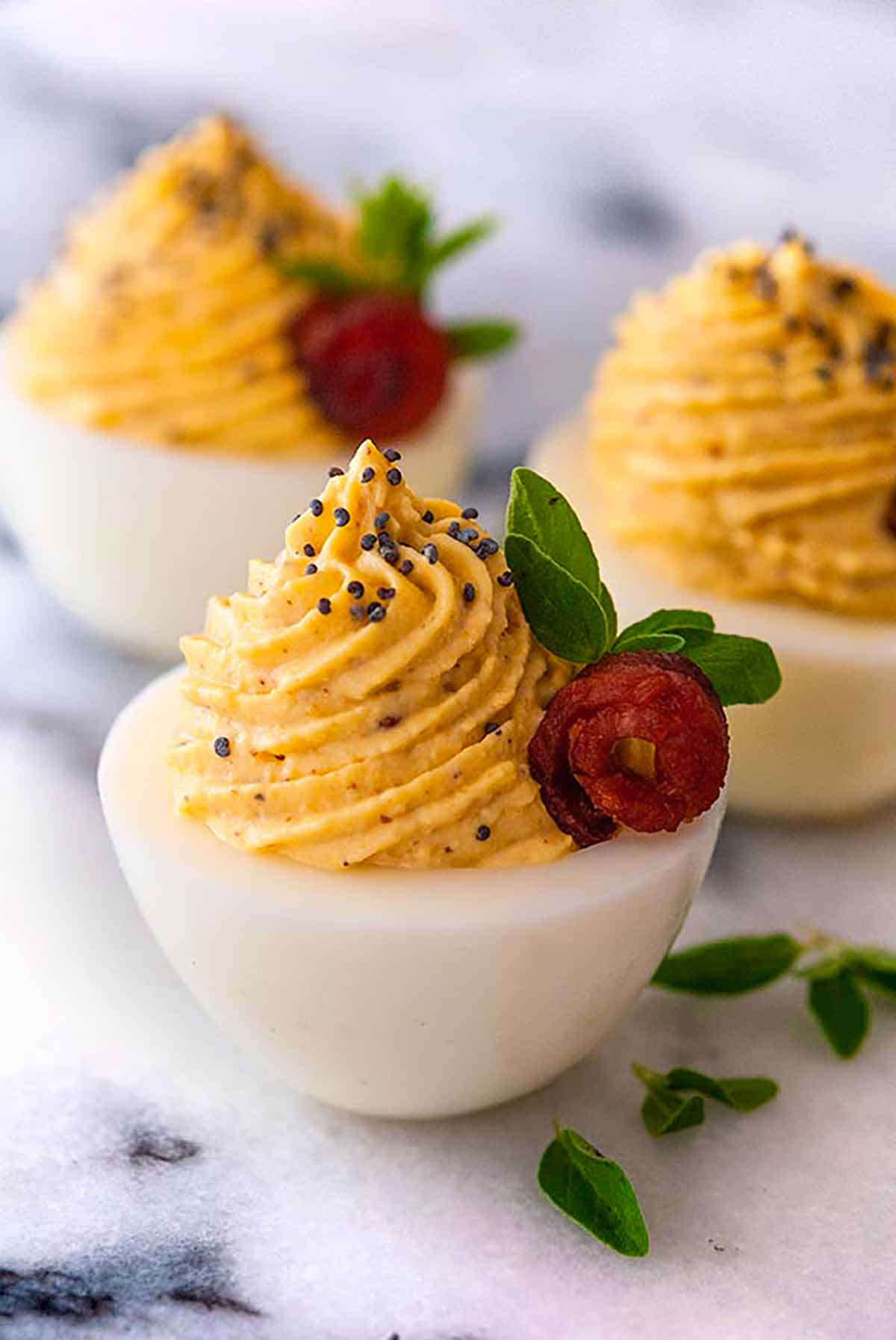 3 deviled eggs garnished with tiny bacon roses, oregano leaves and a sprinkle of poppy seeds.