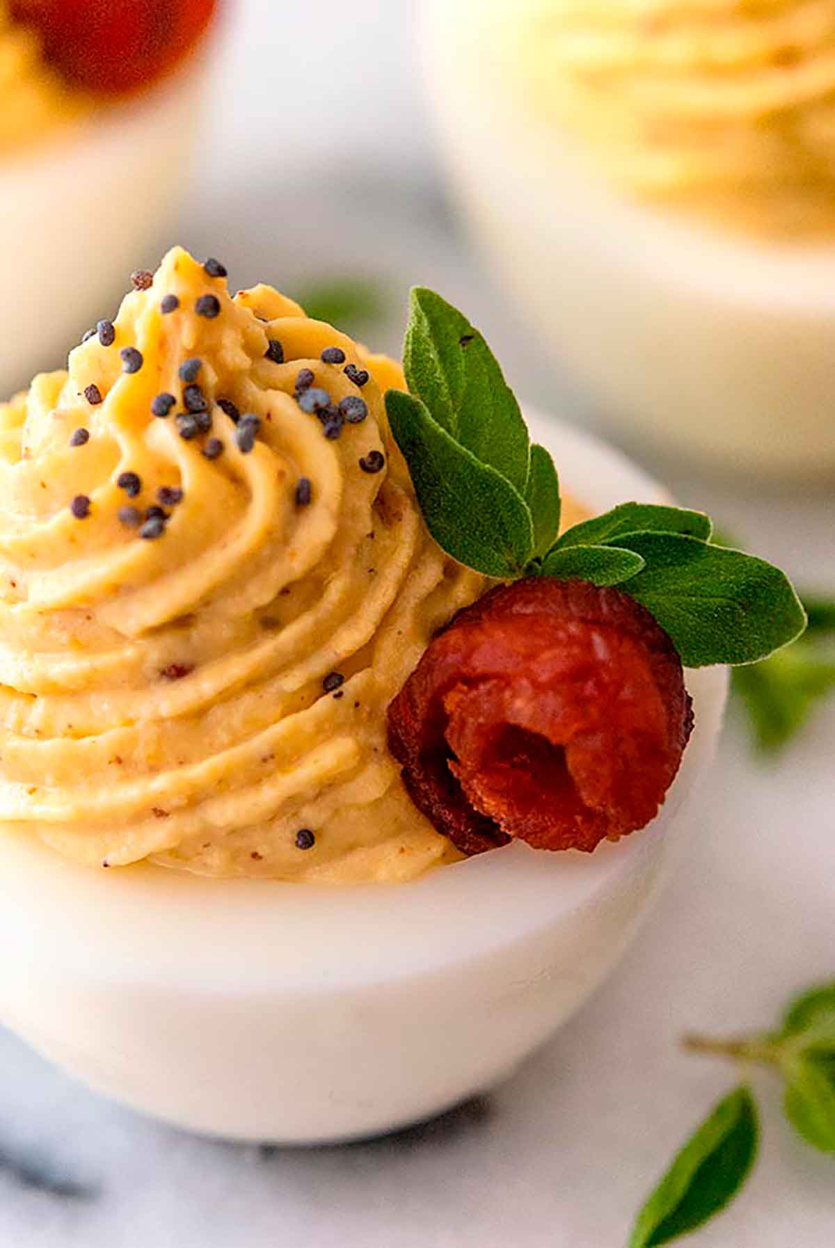 A deviled egg garnished with poppyseeds, a bacon rose, and small leaves on a white plate, surrounded by a few others.