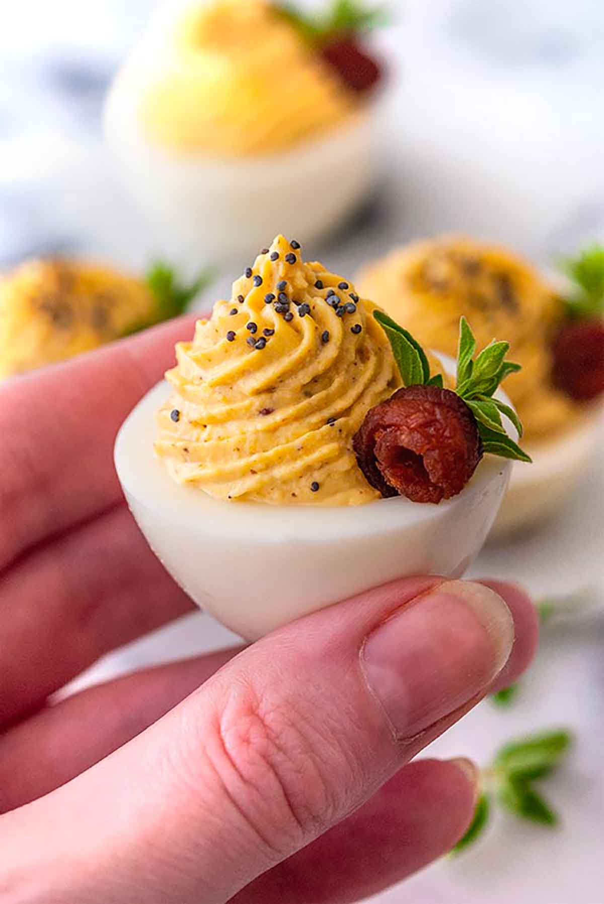 A hand holding a deviled egg, garnished with a bacon rose and oregano leaves.