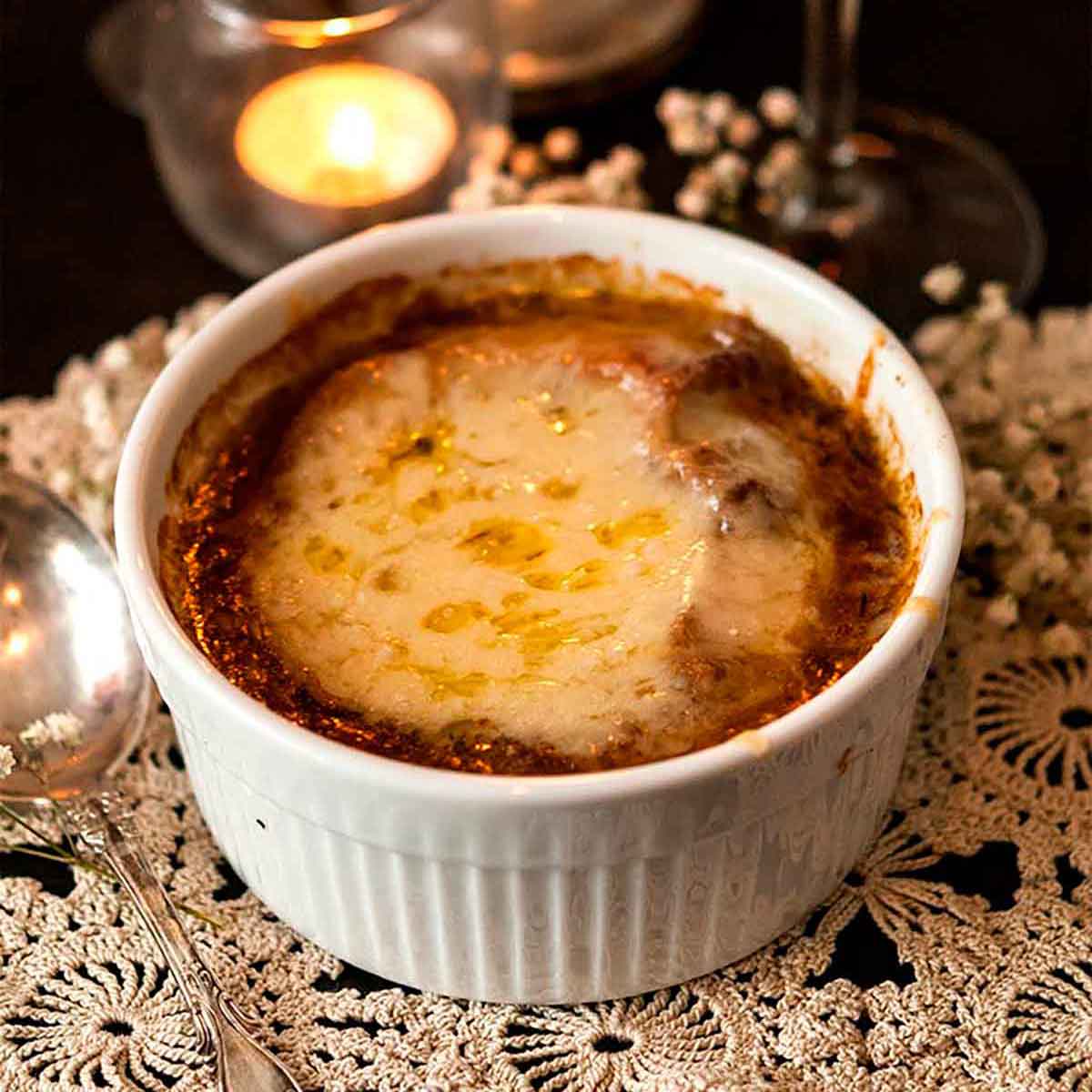 A bowl of French onion soup on a small lace doily in front of a candle, candle stick and glass of red wine.