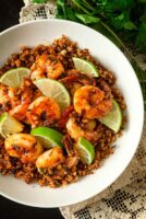 A bowl of lime shrimp atop coconut cauliflower rice on a lace tablecloth with cilantro beside it.