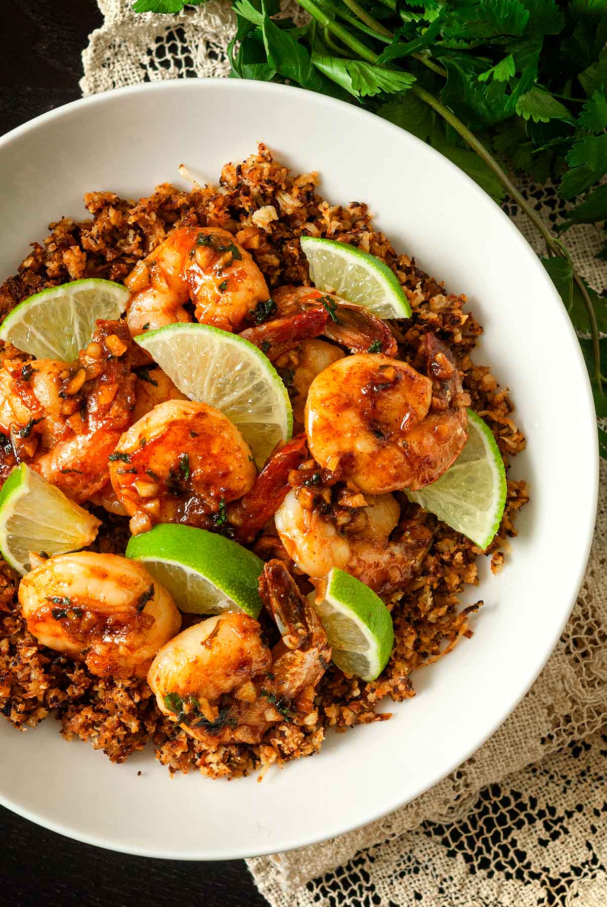 A bowl of lime shrimp atop coconut cauliflower rice on a lace tablecloth with cilantro beside it.