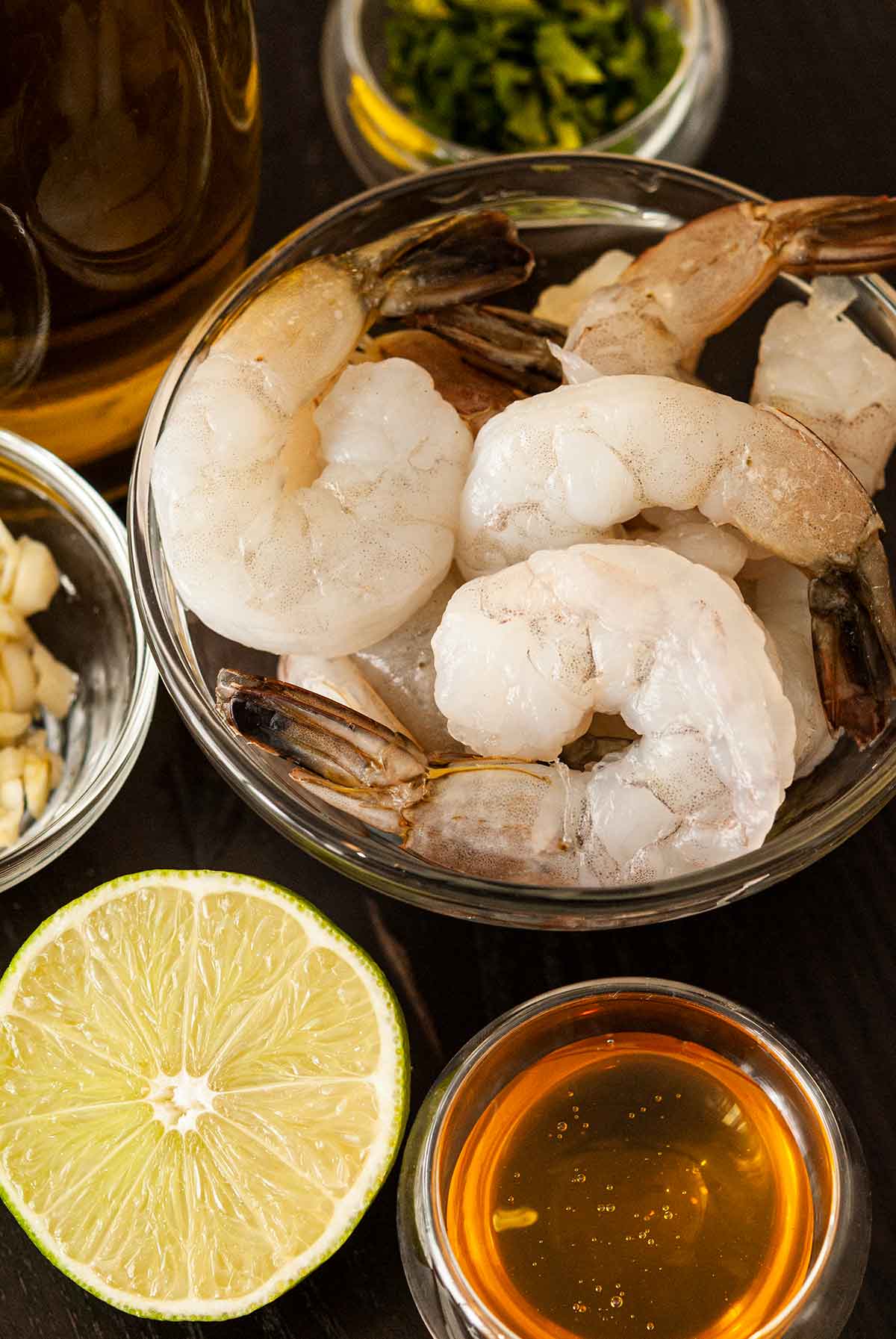 A bowl of raw shrimp next to a sliced lime, surrounded by small bowls with honey, cilantro and garlic.