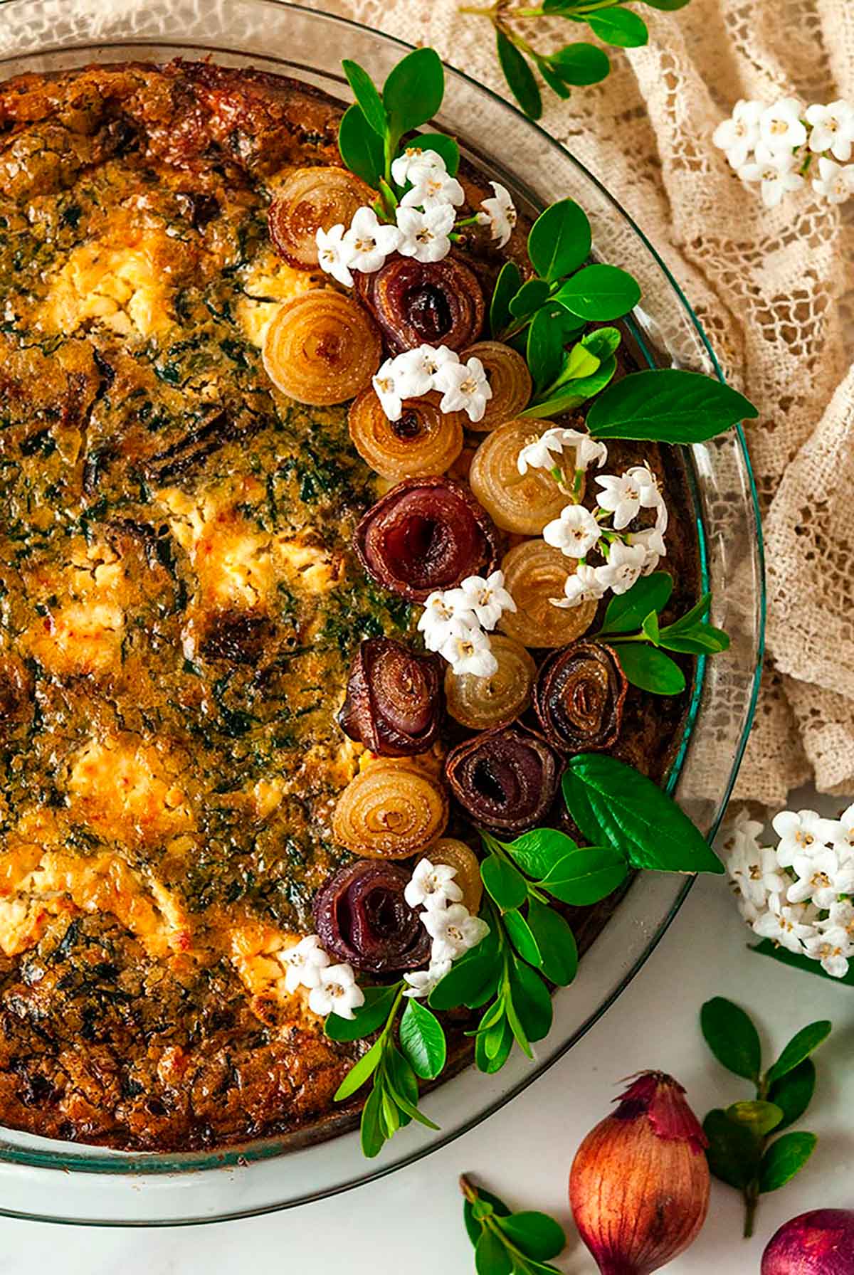 A quiche, garnished with pearl onion roses and little flowers on a marble table with a lace table cloth.