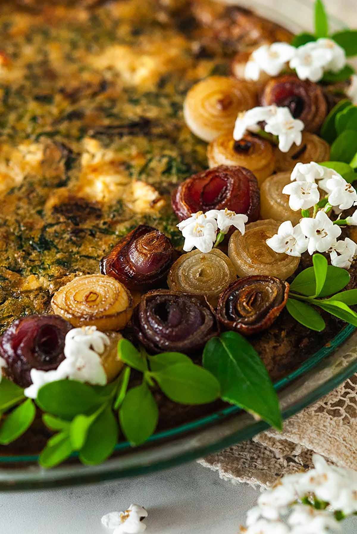 A quiche, garnished with pearl onion roses and little flowers on a marble table with a lace table cloth.