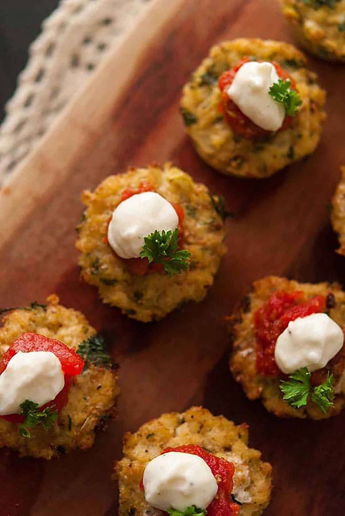 5 cauliflower cakes on a wooden board, topped with marinara, cream, and a little fresh parsley.