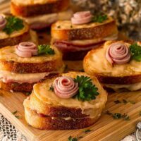 6 mini croque monsieur with a small ham rose garnishes and parsley on a wooden cutting board, surrounded by flowers.