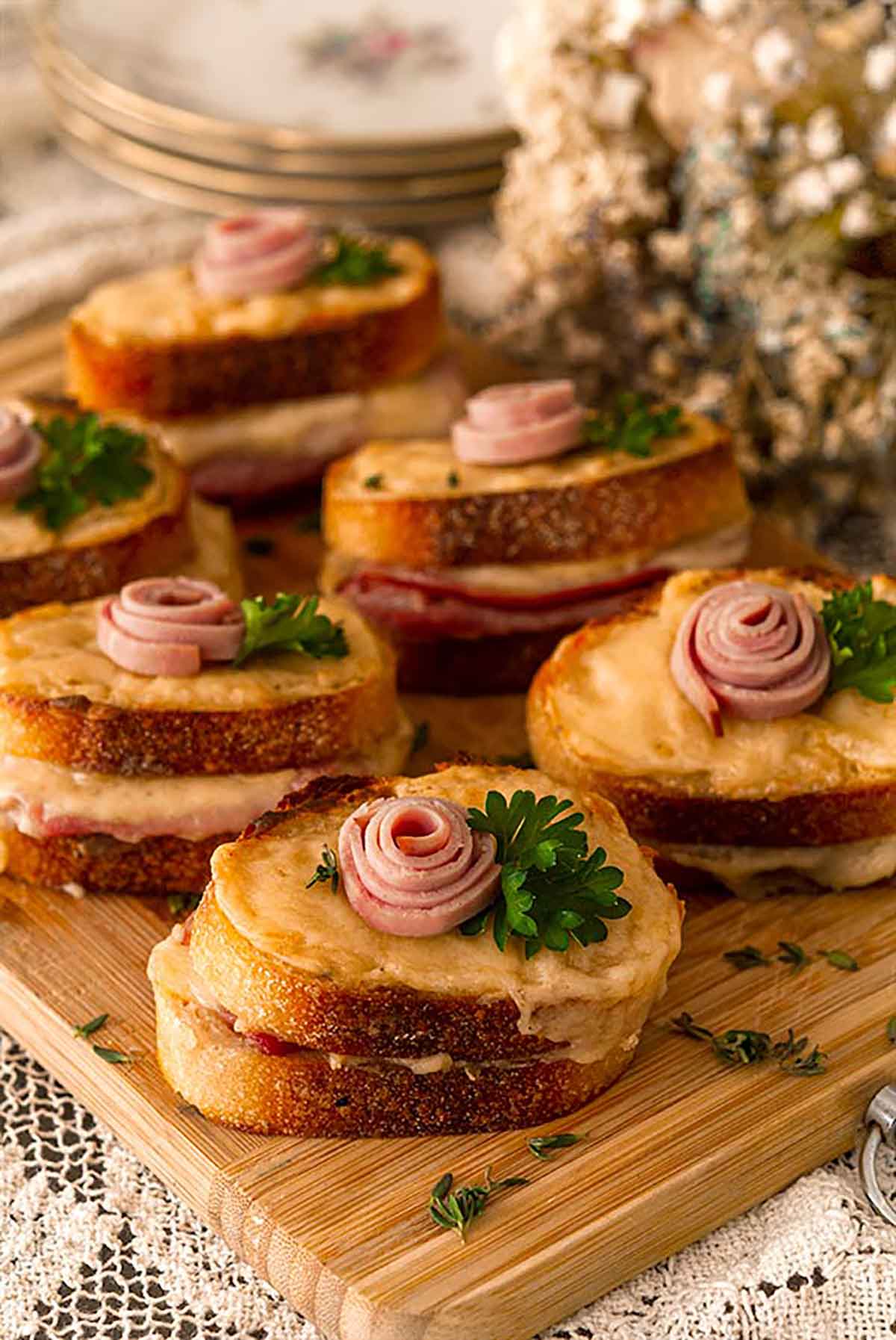 6 mini croque monsieur with a small ham rose garnishes and parsley on a wooden cutting board, surrounded by flowers.