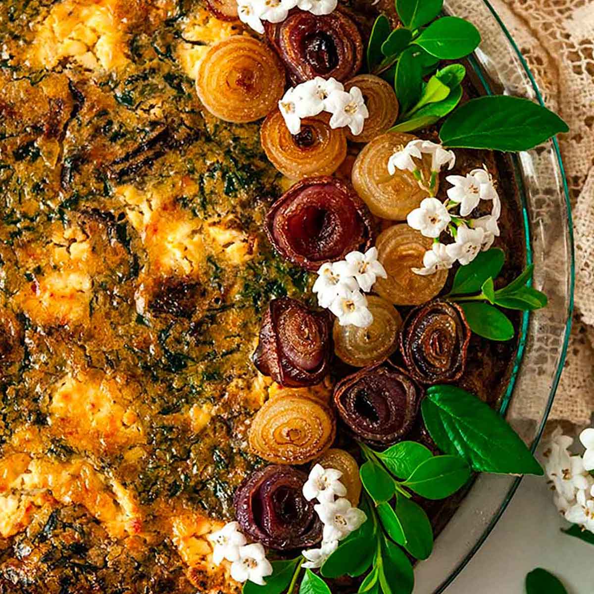 A quiche, garnished with pearl onion roses and little flowers on a marble table with a lace table cloth.