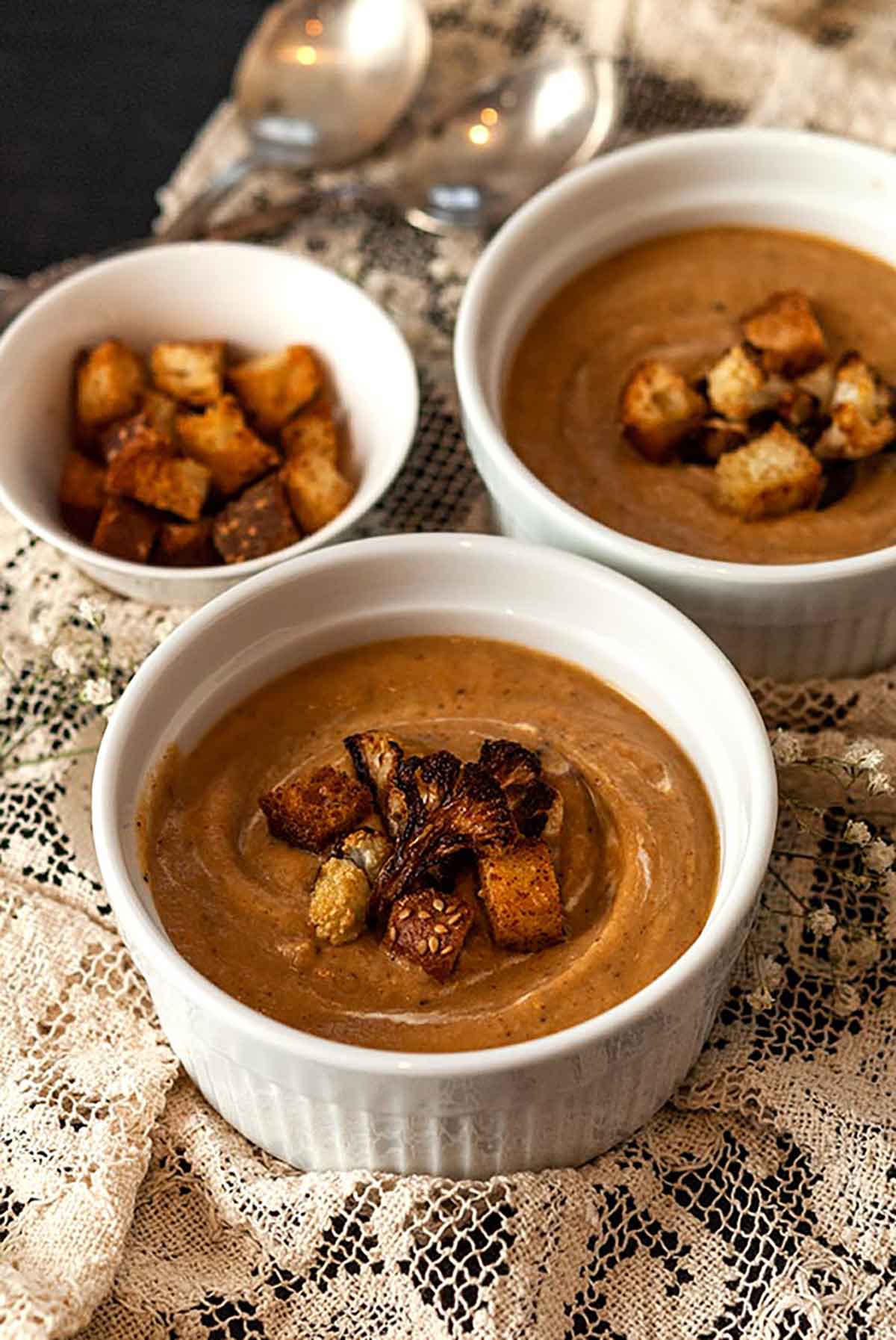 2 bowls of cauliflower soup and a small bowl of croutons on a lace table cloth.