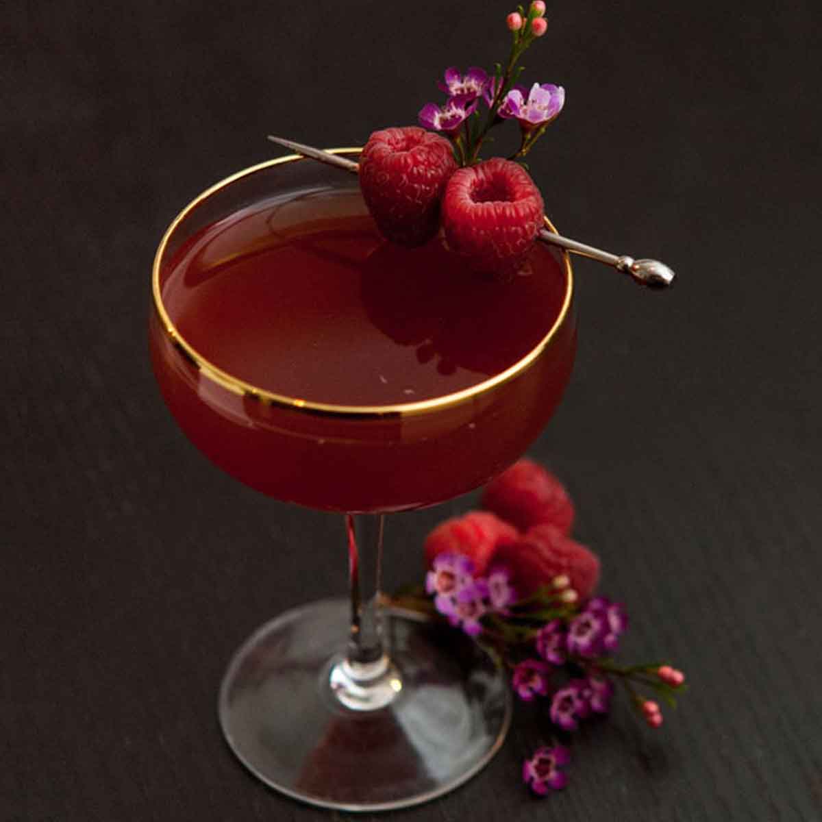 A cocktail on a dark table garnished with raspberries and small flowers, with a few raspberries and flowers at its base.