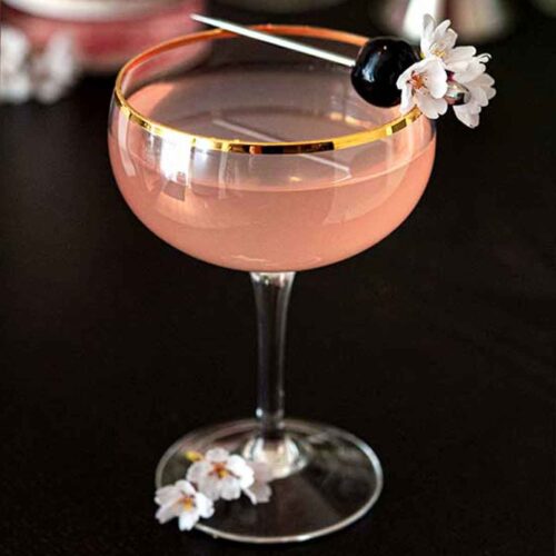 A pink cocktail, garnished with a cherry and small cherry blossoms on a black table.