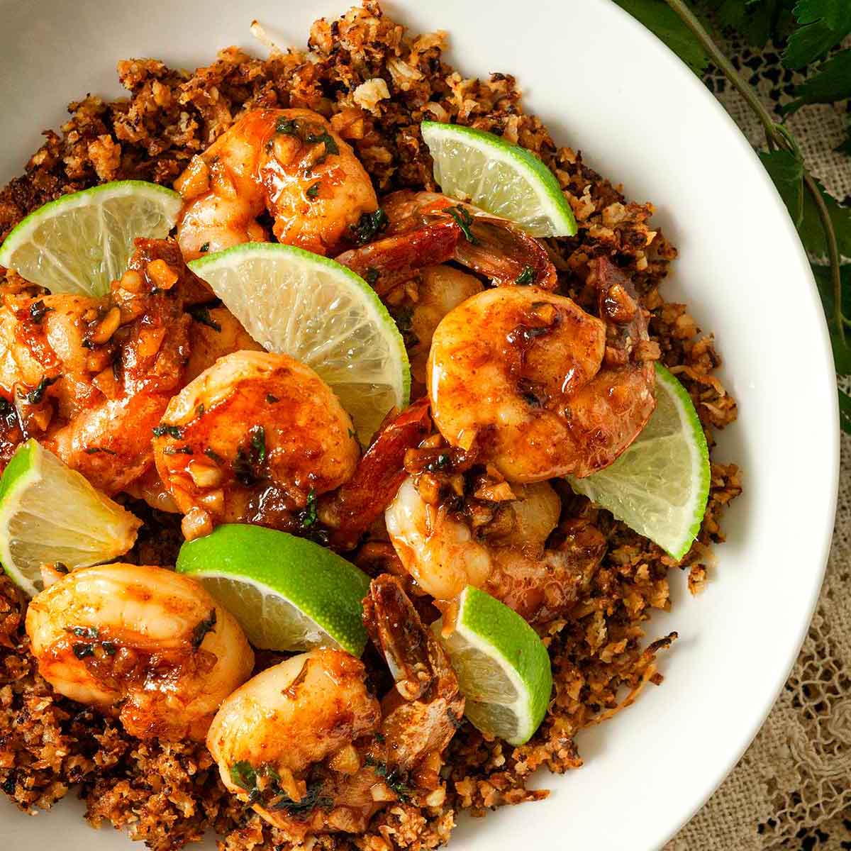 A bowl of lime shrimp atop coconut cauliflower rice on a lace tablecloth with cilantro beside it.