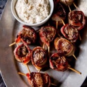 Filet mignon bites, wrapped in bacon and stuck with toothpicks, beside a sauce on a metal plate.