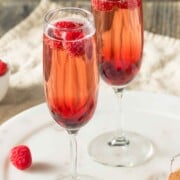 2 Kir Royale cocktails in champagne flutes on a table, garnished with raspberries.