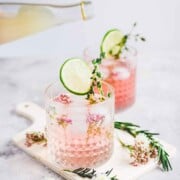 2 pink cocktails on a white table, garnished with thyme and lime slices.