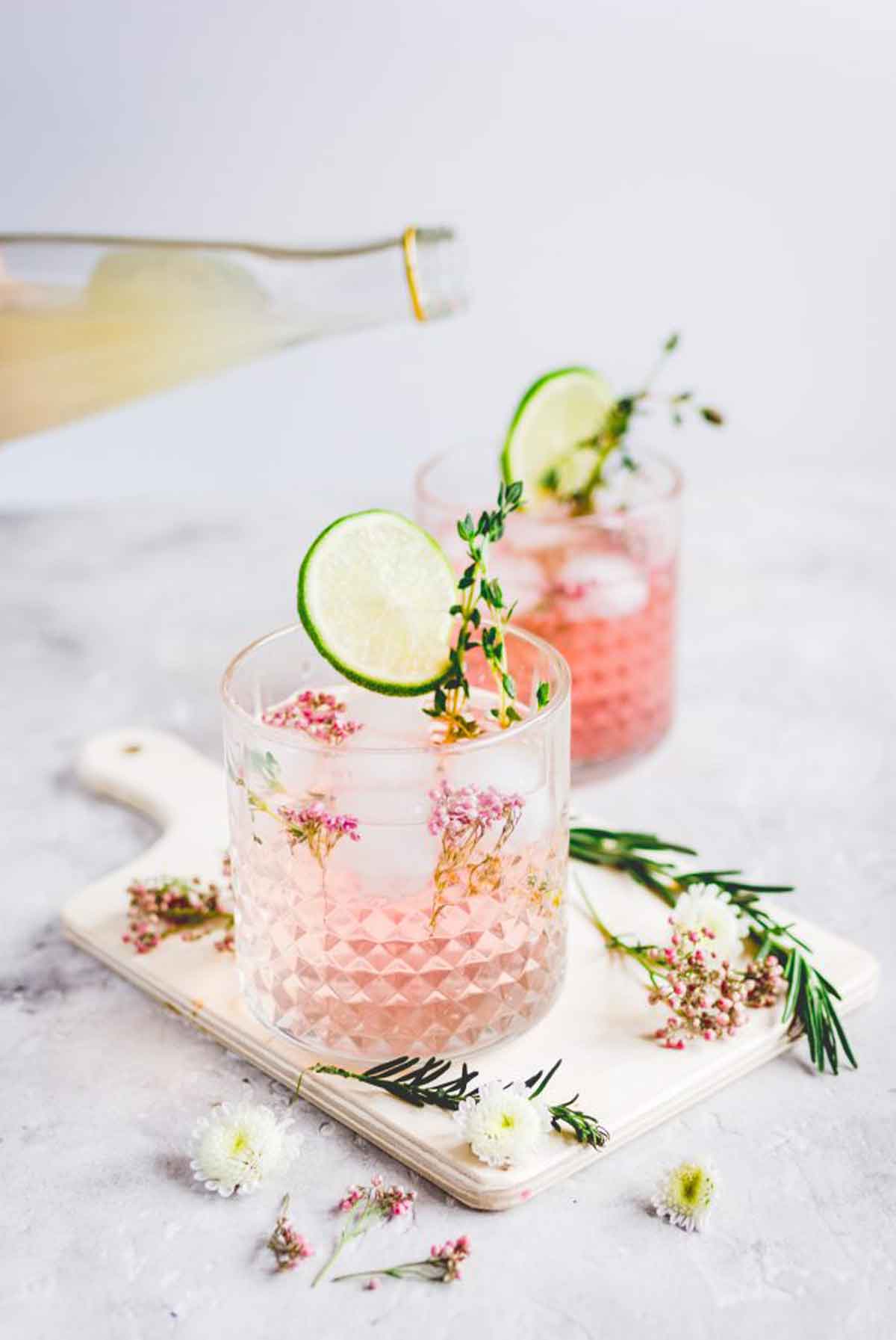 2 pink cocktails on a white table, garnished with thyme and lime slices.
