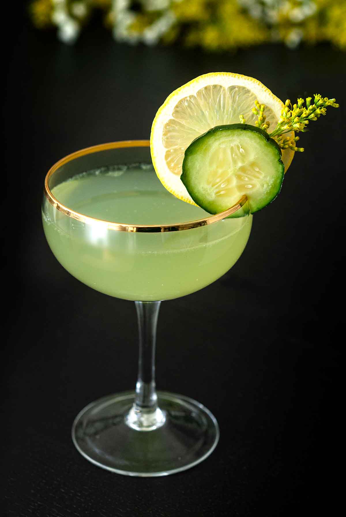 A cocktail in a coup glass, garnished with a cucumber slice, lemon, and wispy flower on a table.