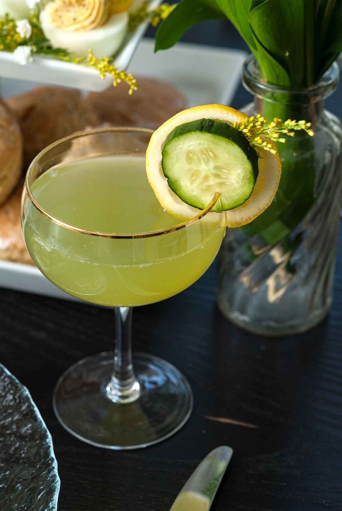 A cocktail on a table in front of brunch food and a small vase of greenery on a table.