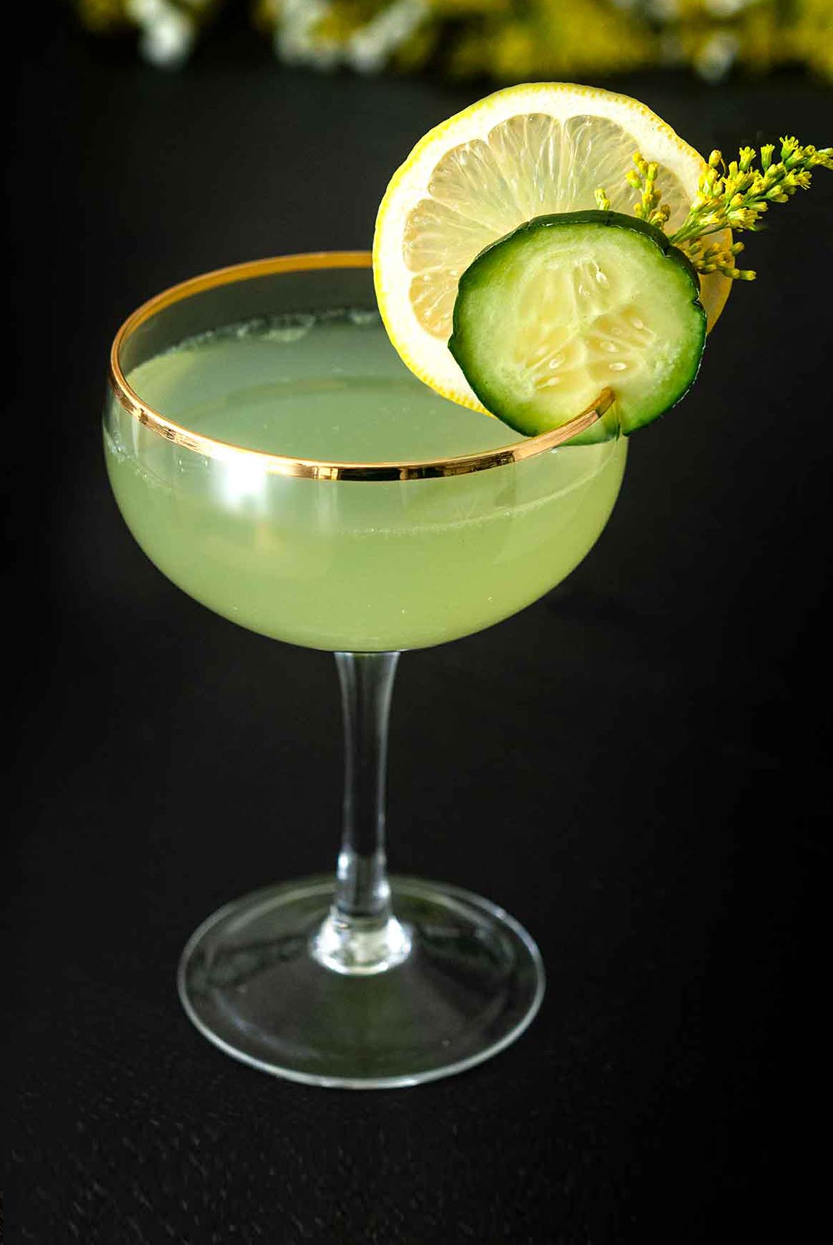 A cocktail in a coup glass, garnished with a cucumber slice, lemon, and wispy flower on a table.