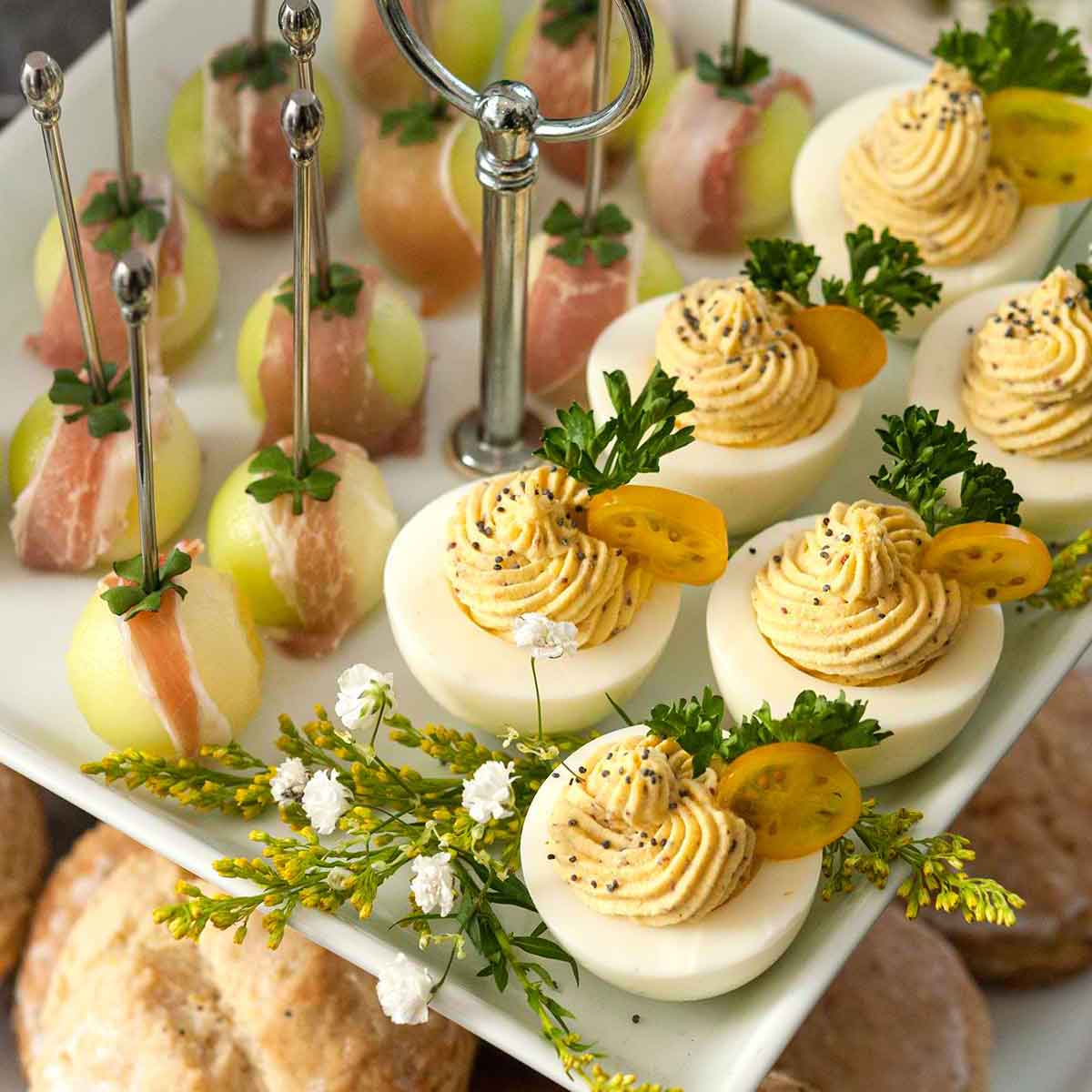 An appetizer tier with deviled eggs and prosciutto-wrapped melon above scones on a table.