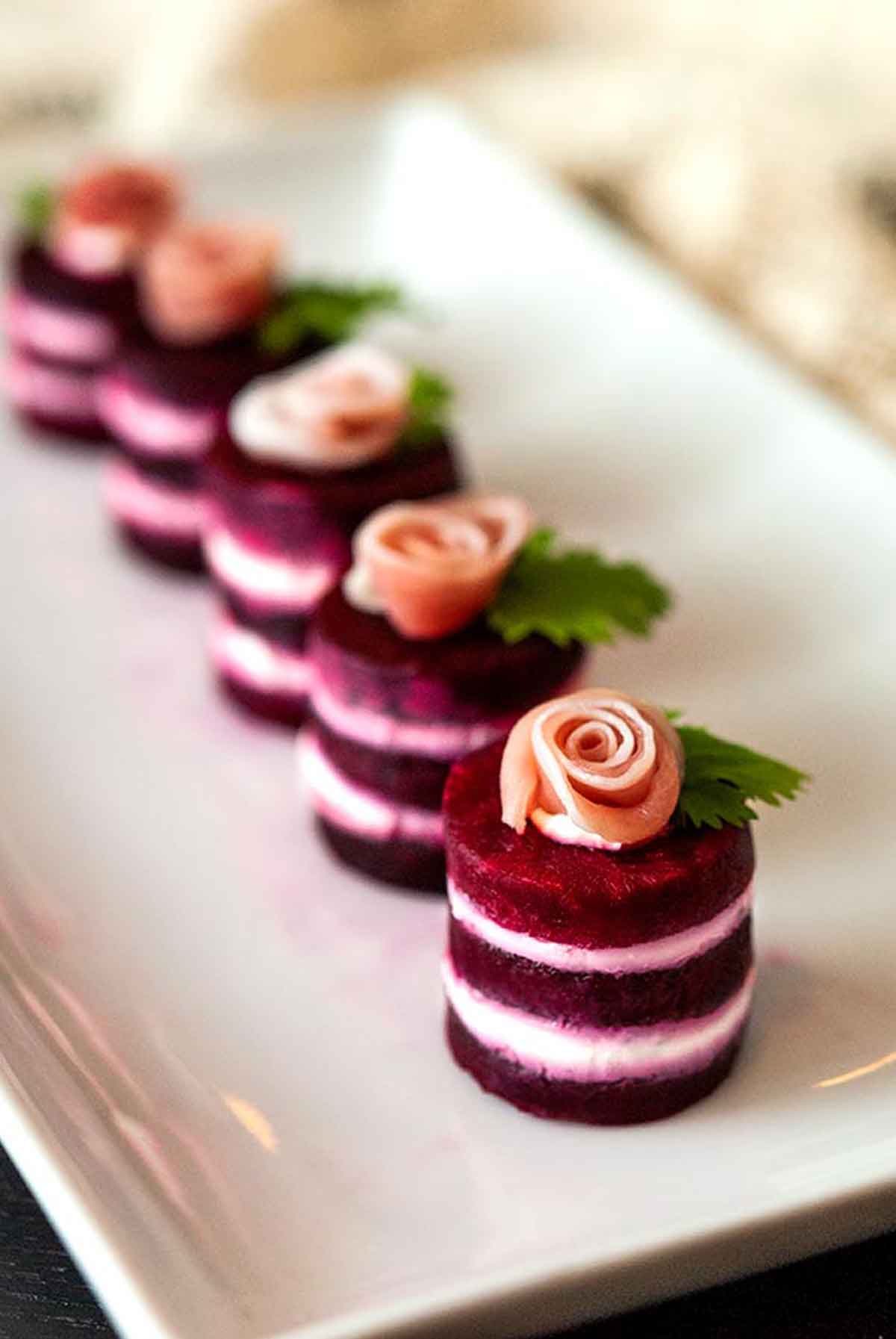 6 beet napoleons, with prosciutto roses on top, garnished with cilantro leaves on a plate.