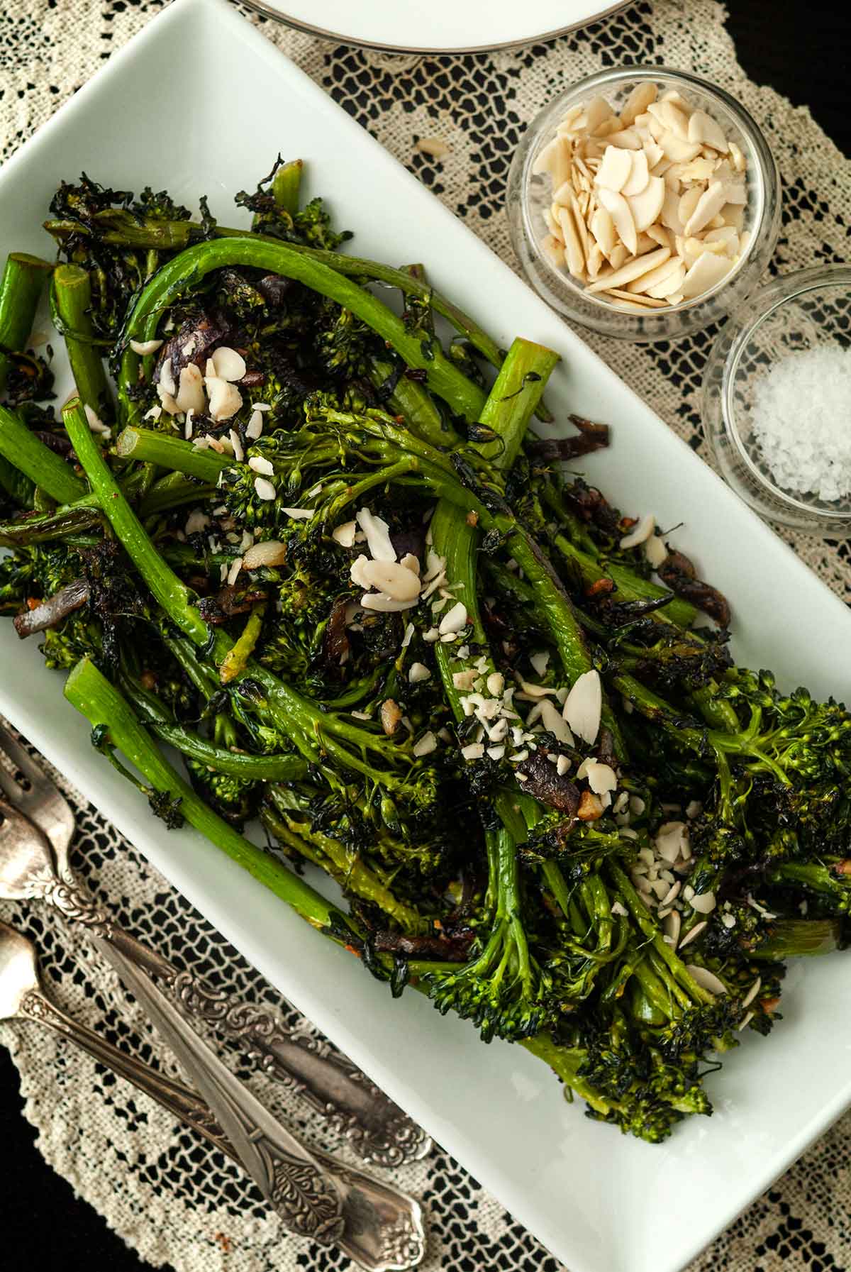 A plate of sautéed broccoli rabe on a lace tablecloth, beside 2 small bowls of garnish and 2 antique forks.