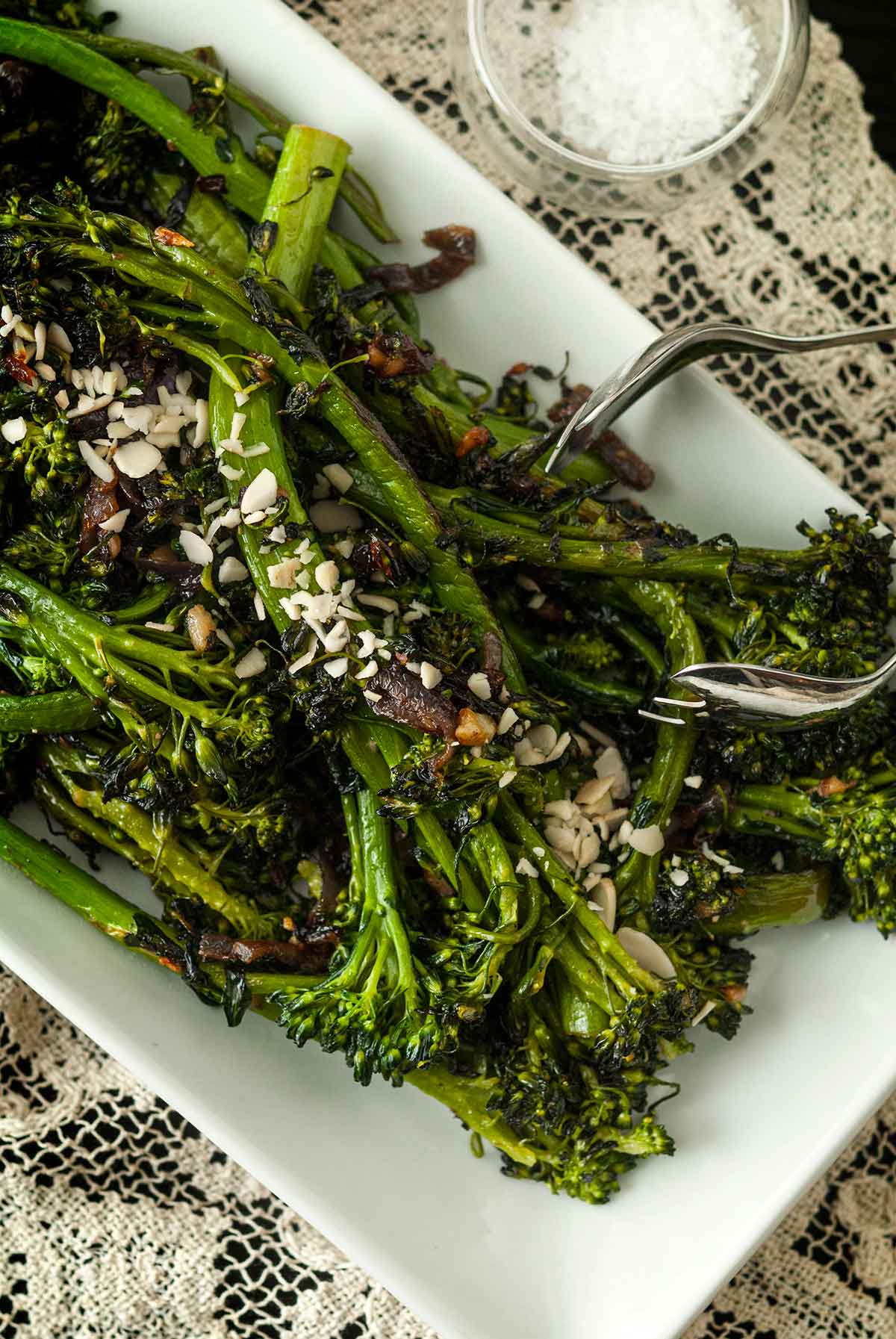 A plate of sautéed broccoli rabe on a lace tablecloth, beside a small bowl of salt, with salad tongs.