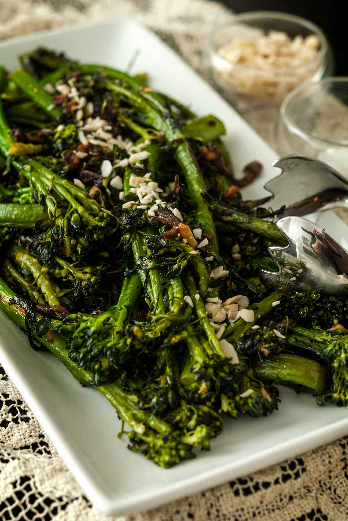 A plate of sautéed broccoli rabe on a lace tablecloth, beside 2 small bowls of garnish, with salad tongs.