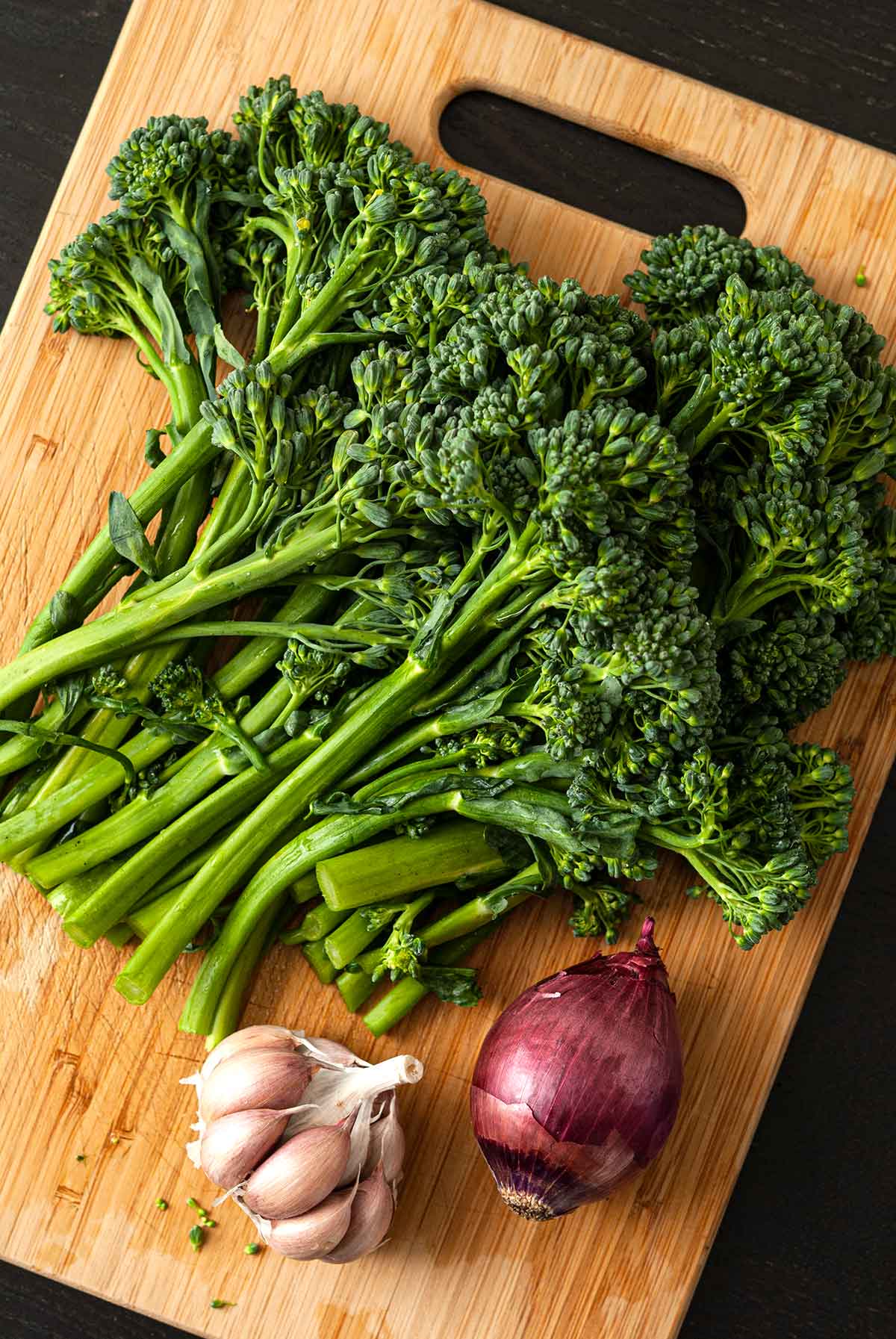 A bunch of raw broccoli rabe on a wooden cutting board with a small red onion and head of garlic.