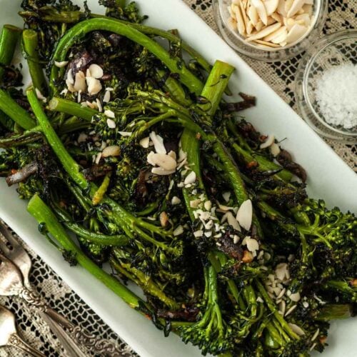 A plate of sautéed broccoli rabe on a lace tablecloth, beside 2 small bowls of garnish and 2 antique forks.