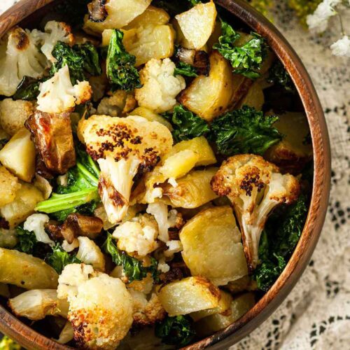 A wooden bowl of potato-cauliflower hash on a lace table cloth with flowers on the edges.