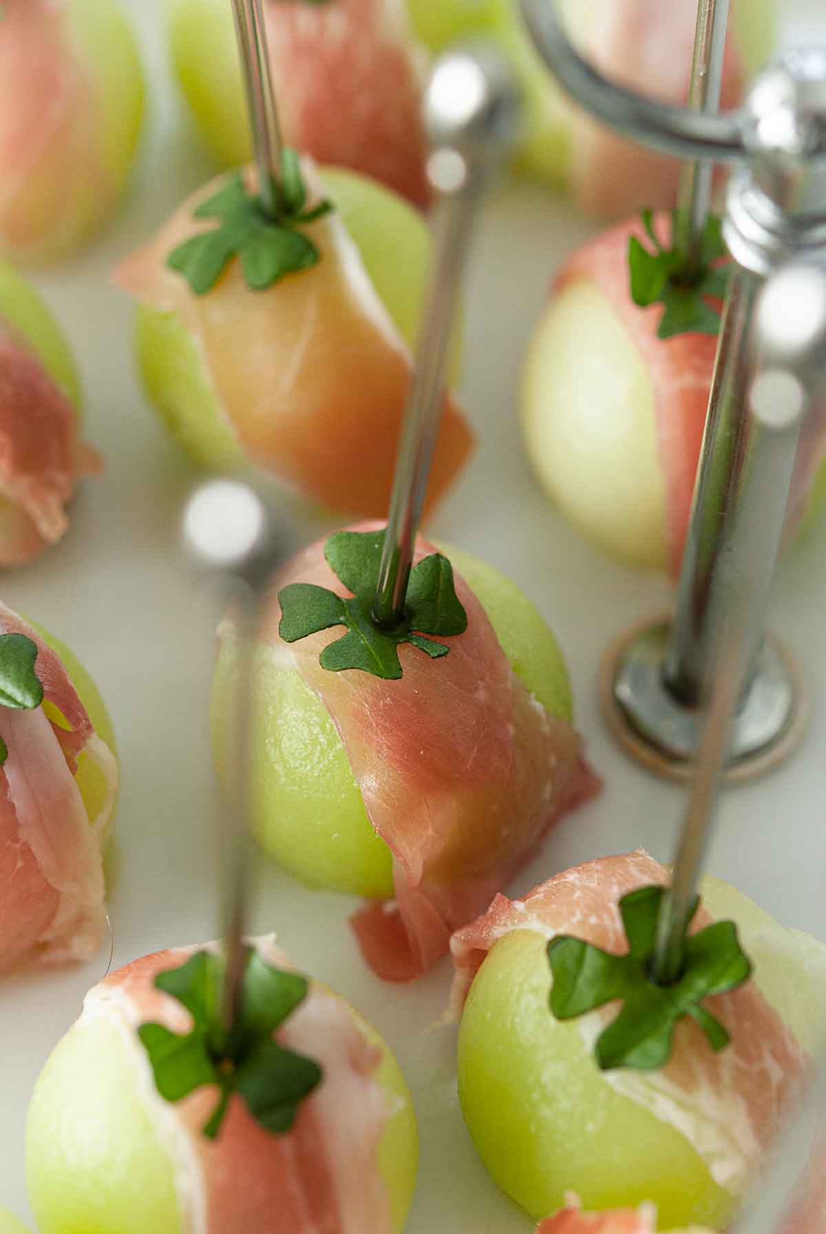 About 10 balled melon appetizers, wrapped in prosciutto with clovers on top, and pierced with cocktail pins.