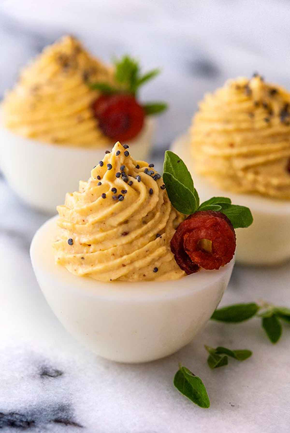 3 deviled eggs garnished with tiny bacon roses, oregano leaves and a sprinkle of poppy seeds.