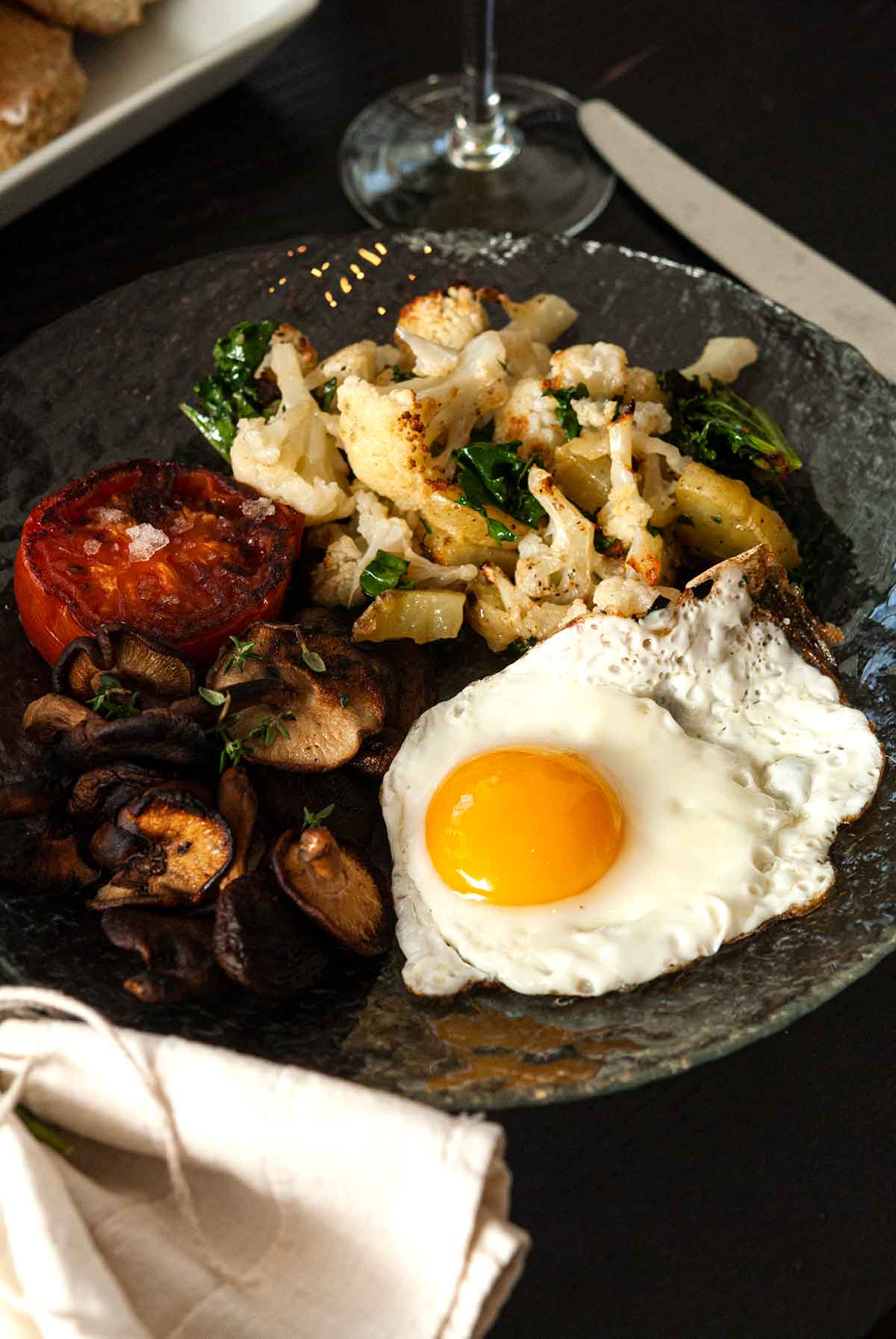 A plate of potato-cauliflower hash, a tomato, mushrooms and 1 egg on a table.