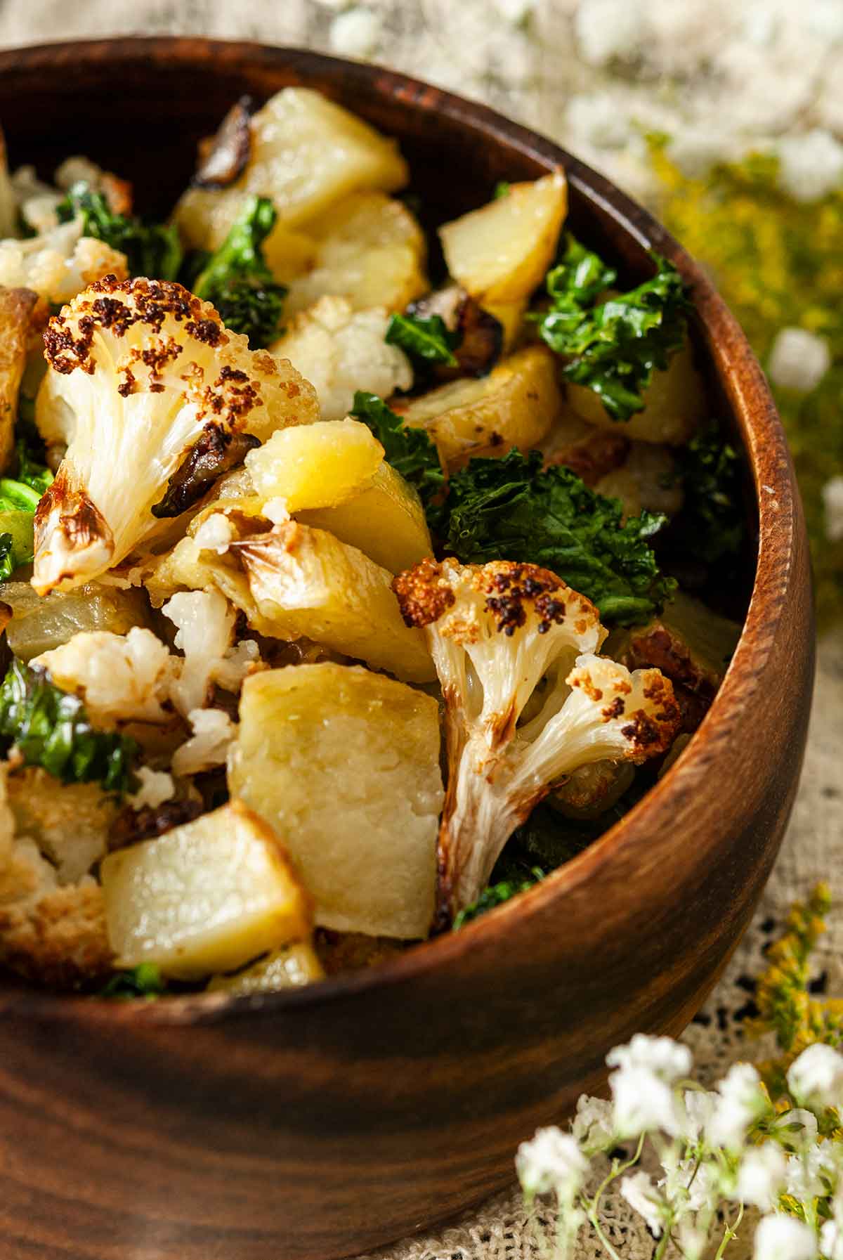 A close photo of a wooden bowl of potato-cauliflower hash on a lace table cloth with flowers on the edges.