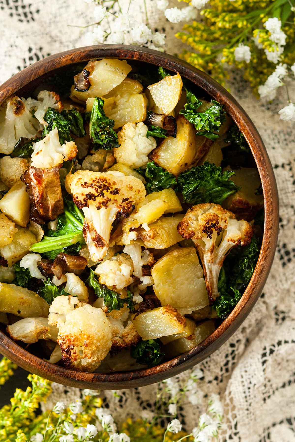 A wooden bowl of potato-cauliflower hash on a lace table cloth with flowers on the edges.