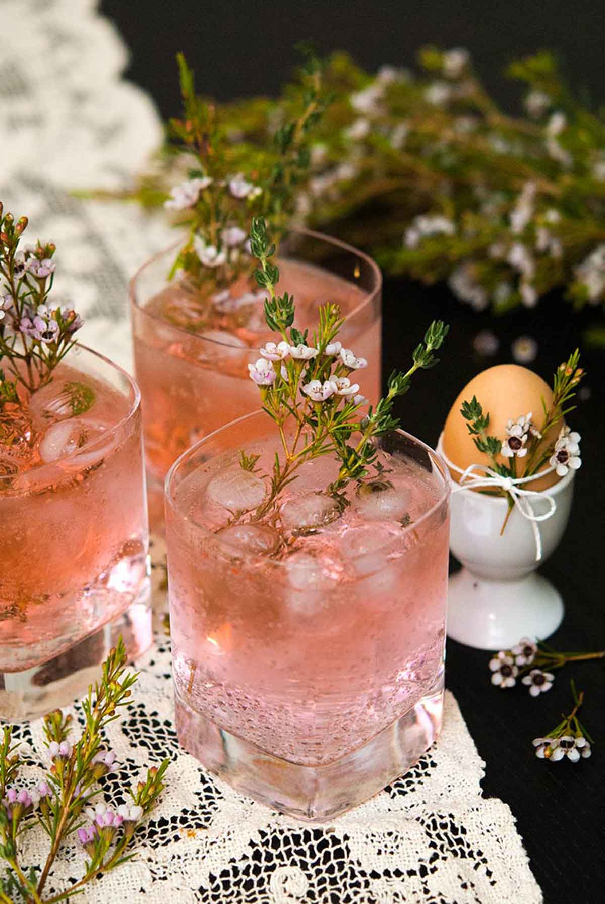 3 pink gin and tonic cocktails on a lace table cloth garnished with and surrounded by floral greenery and thyme, beside an egg in an egg cup.