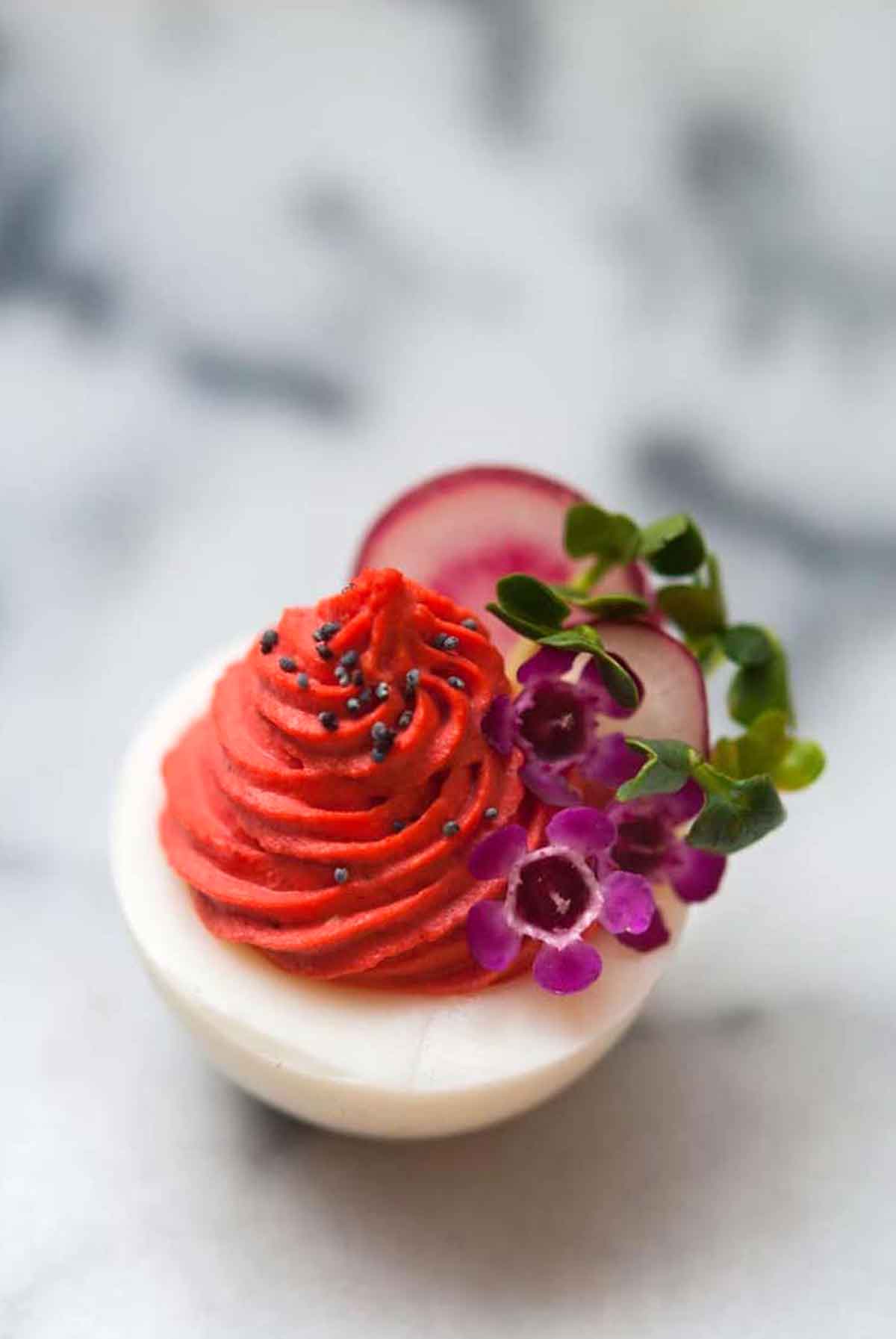 A red deviled egg, garnished with flowers and radishes on a marble slate.