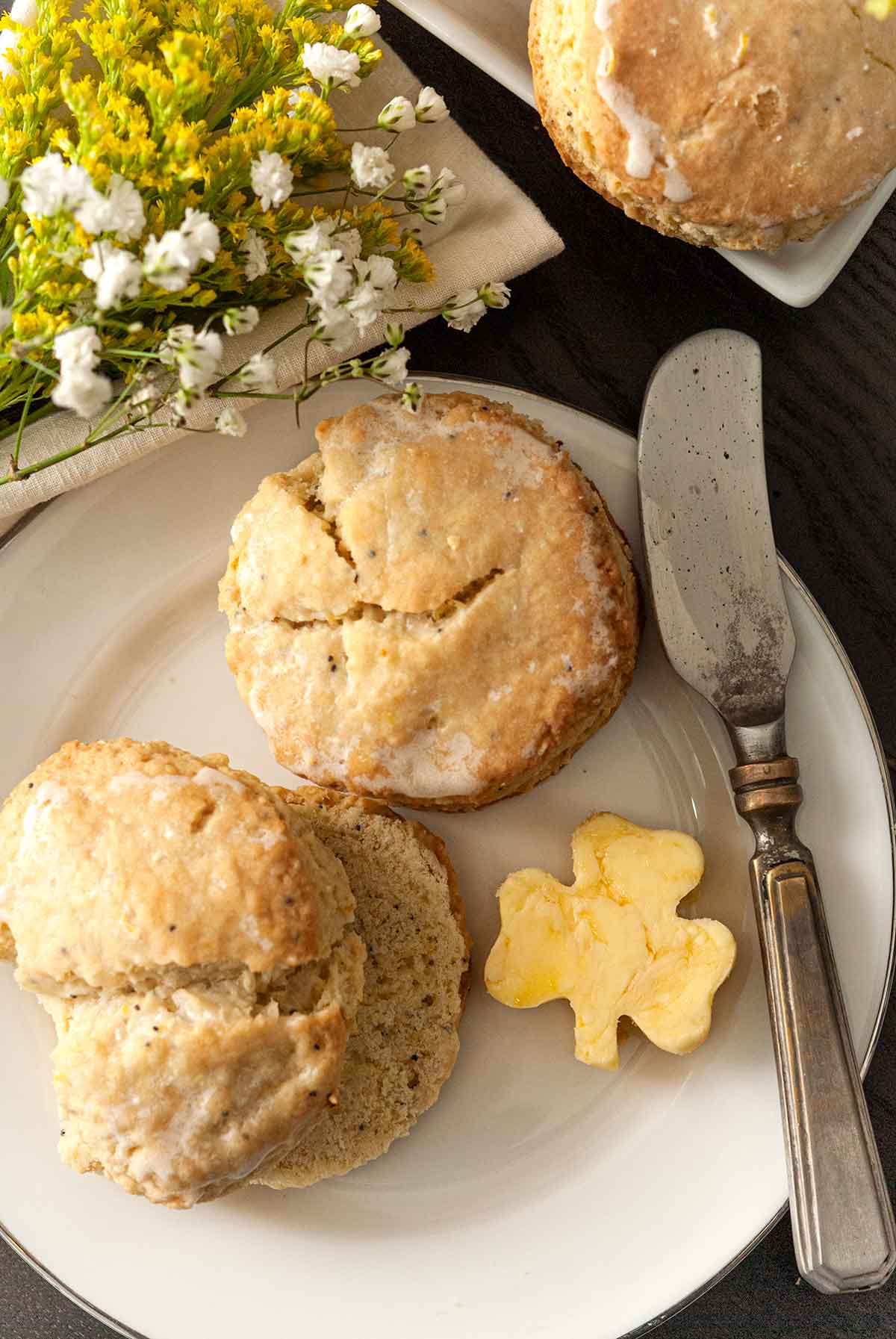 2 scones on a plate with a shamrock-shaped pad of butter, beside a knife, in front of a few flowers o a table.