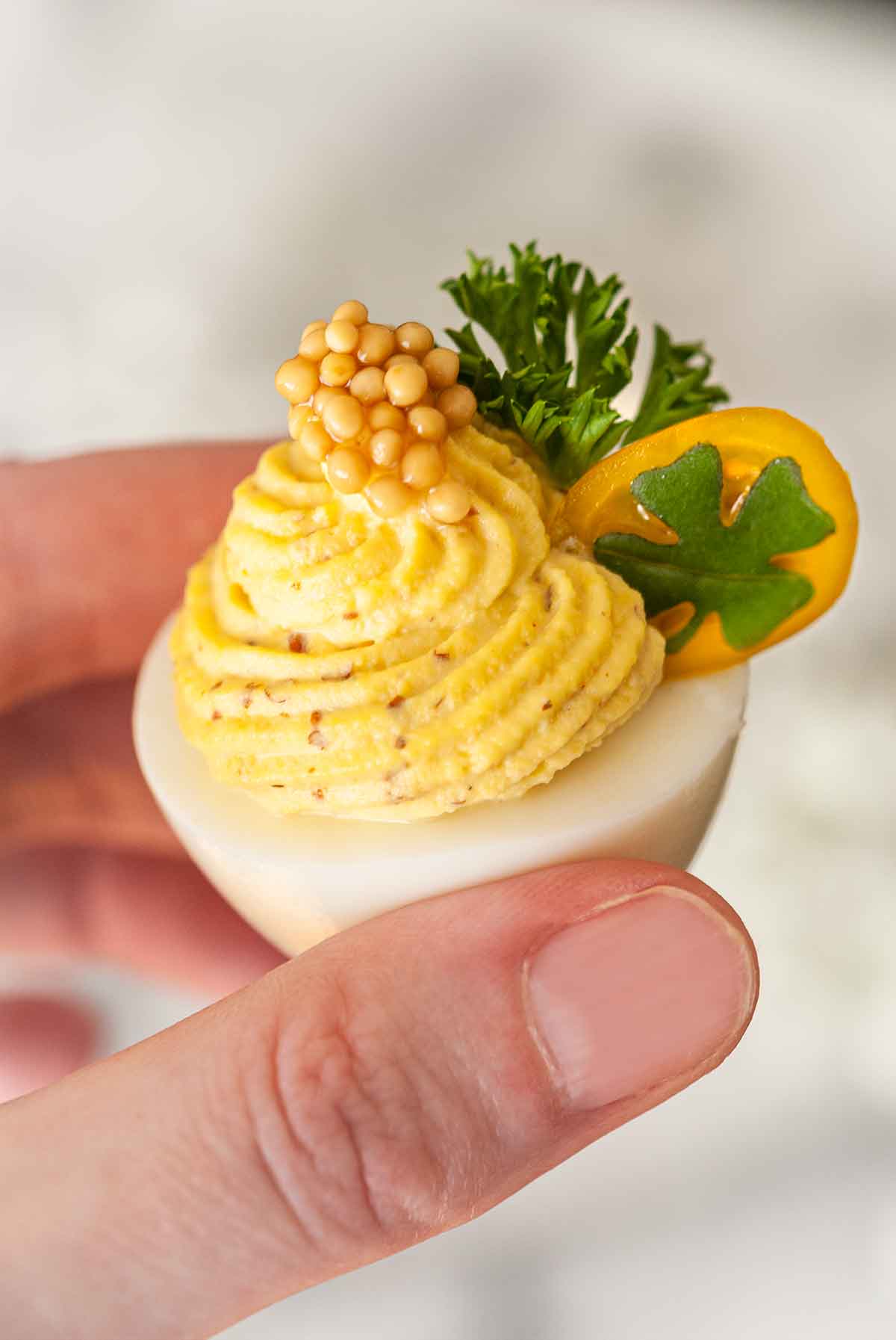 A hand holding a deviled egg, garnished with tomato, clover, parsley and a dollop of mustard caviar.