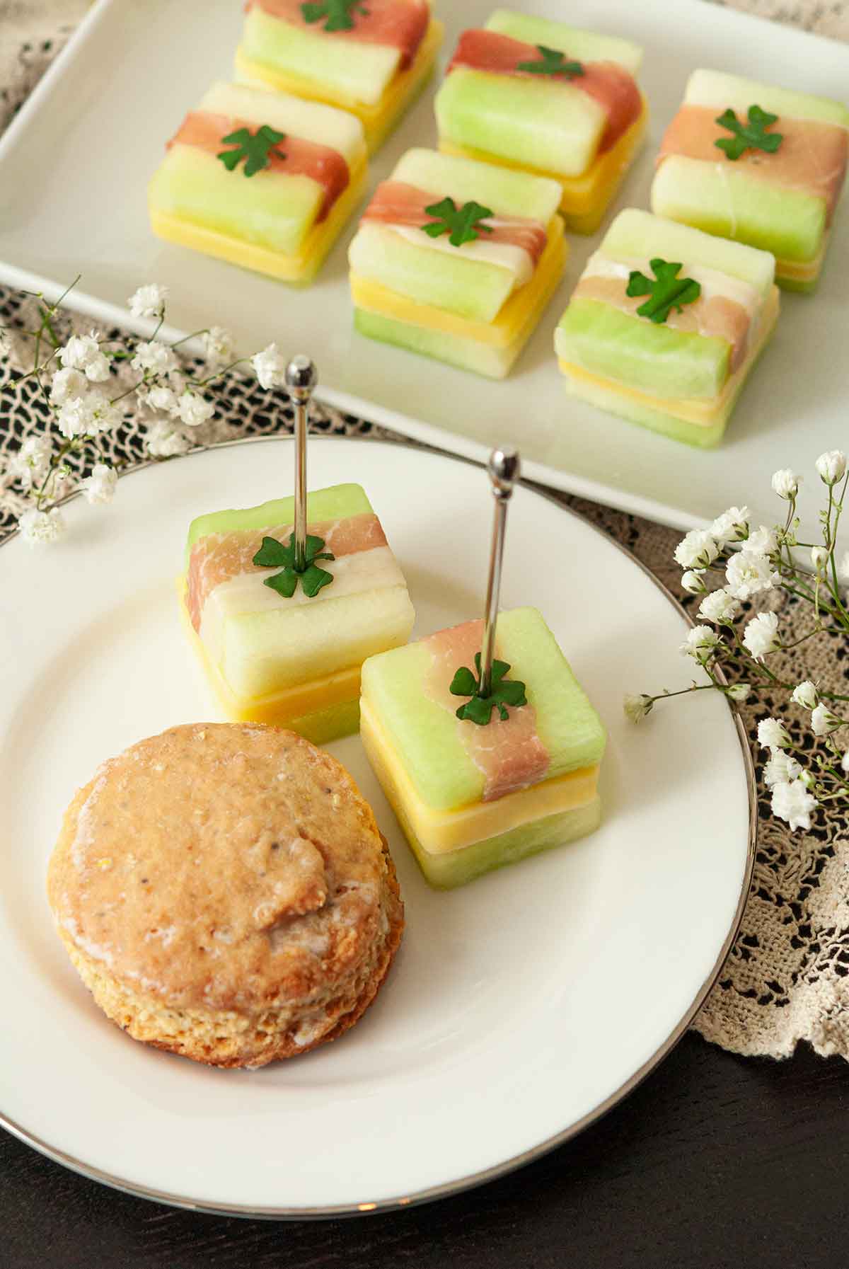 2 melon appetizers on a plate with a scone in front of 6 more melon appetizers on a plate.